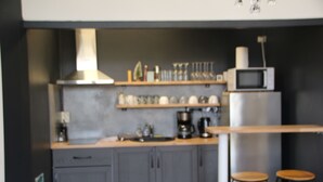 A kitchen with grey cabinets, a microwave, a coffee maker, and two stools. - Le km99 (Pont-sur-Yonne)