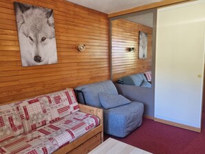 Une pièce avec un mur en bois, un canapé recouvert d’un tissu à motifs, un fauteuil gris et une porte avec un miroir.