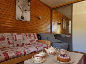 A cozy room with a wooden wall, a patterned sofa, a blue armchair, and a table with a teapot, cups, and a cake.