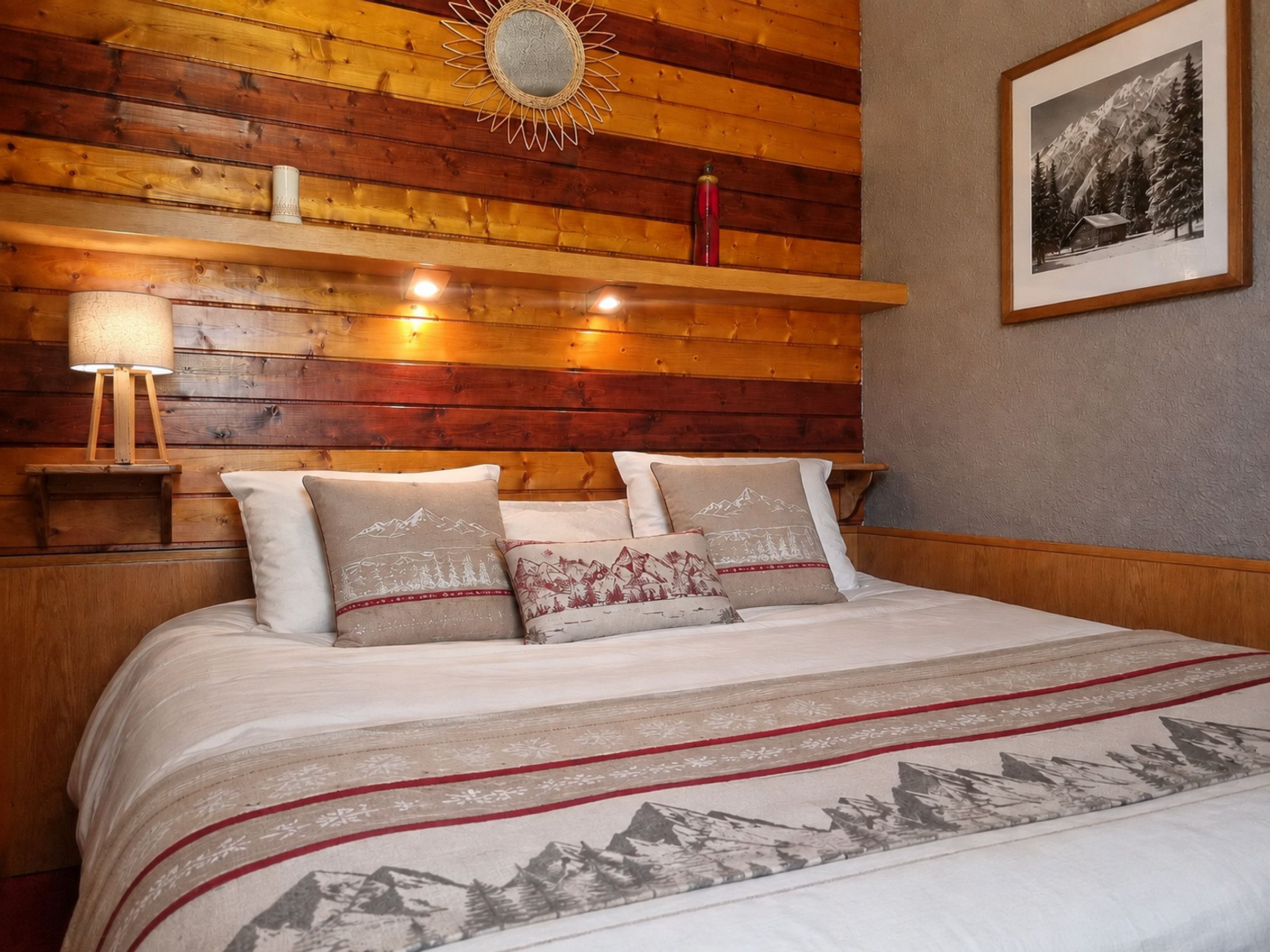 Ein Bett mit einem zudeckenden Bettbezug mit Bergmotiv, einem Holzkopfbrett und Nachttischlampen.