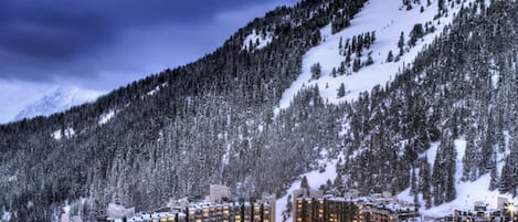 A snow-covered resort with multiple high-rise buildings, ski lifts, and a mountainous backdrop.
