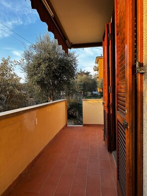 A balcony with red tiles, a wooden door, and a view of trees and a building in the background. - Casa Gabriel (Toscolano Maderno)