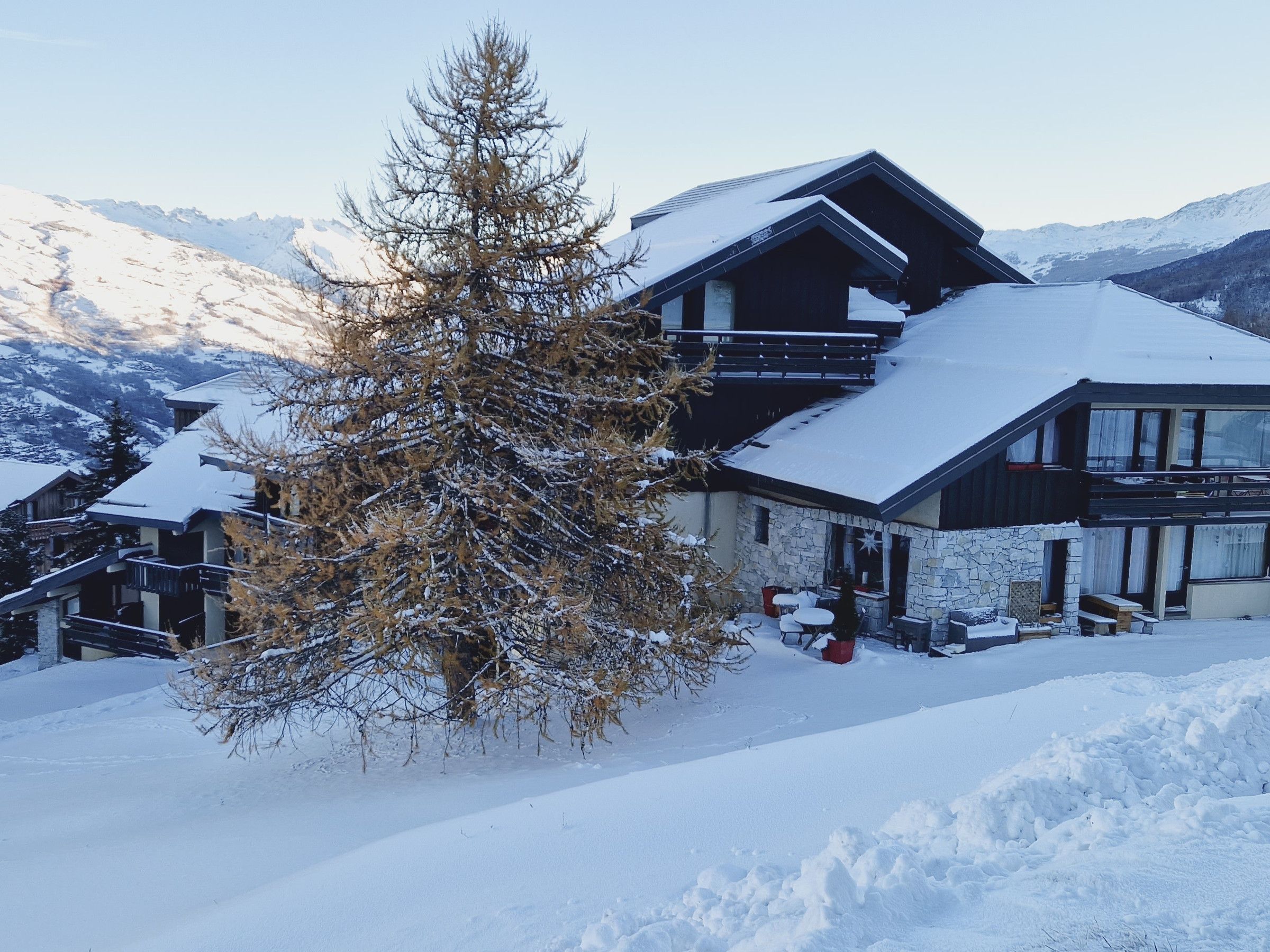 A two-story chalet with a stone facade, wooden balconies, and a snow-covered roof.