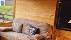 A cozy room with a wooden wall, a cushioned sofa with pillows, a small wooden table, and a window overlooking a grassy area. - Cozy Studio Near Ski Slopes, Ideal for Families, Fully Equipped Kitchen (La Plagne Montalbert)