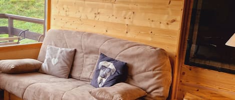A cozy room with a wooden wall, a cushioned sofa with pillows, a small wooden table, and a window overlooking a grassy area.
