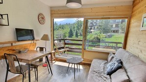 A cozy room with a wooden table, chairs, a sofa, and a large window offering a view of greenery and buildings. - Cozy Studio Near Ski Slopes, Ideal for Families, Fully Equipped Kitchen (La Plagne Montalbert)