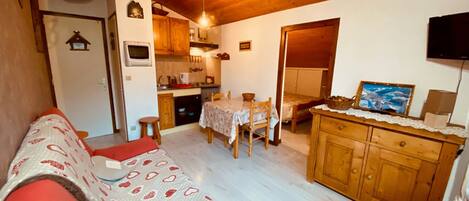 A cozy living room with a red sofa, a wooden dining table, and a kitchen area with wooden cabinets and a television.