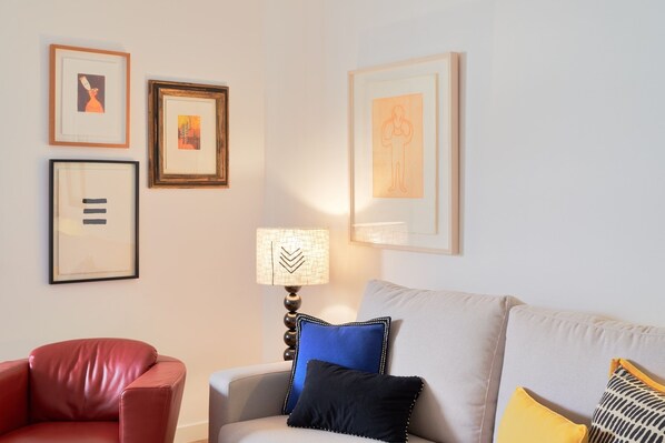 Un salon avec un fauteuil rouge, un canapé gris et une lampe. Des tableaux encadrés ornent le mur.