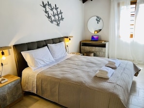 A neatly made bed with a headboard, a bedside table with a lamp, a mirror, and a laptop on a dresser. - Beach Walk Haven: Cozy Home 8 Minutes from Almyrida Beach (Almyrida)