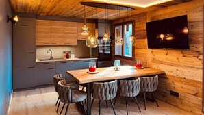 A modern kitchen with wooden walls, a dining area with a wooden table and chairs, pendant lights, and a flat-screen TV mounted on the wall. - Luxury 8-Person Apartment, South Balcony, 120m from Pra-Loup 1500 Lift (Pra-Loup)