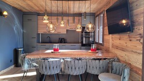 A modern kitchen with wooden accents, pendant lighting, a large island, and a flat-screen TV mounted on the wall. - Luxury 8-Person Apartment, South Balcony, 120m from Pra-Loup 1500 Lift (Pra-Loup)