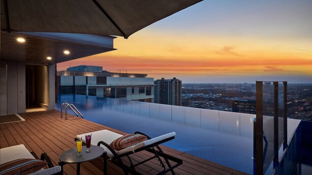 Une piscine à débordement offrant une vue sur la ville au coucher du soleil, des chaises longues et une table avec des boissons.