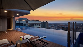 Una piscina infinita con vistas a la ciudad al atardecer, sillas de descanso y una mesa con bebidas.