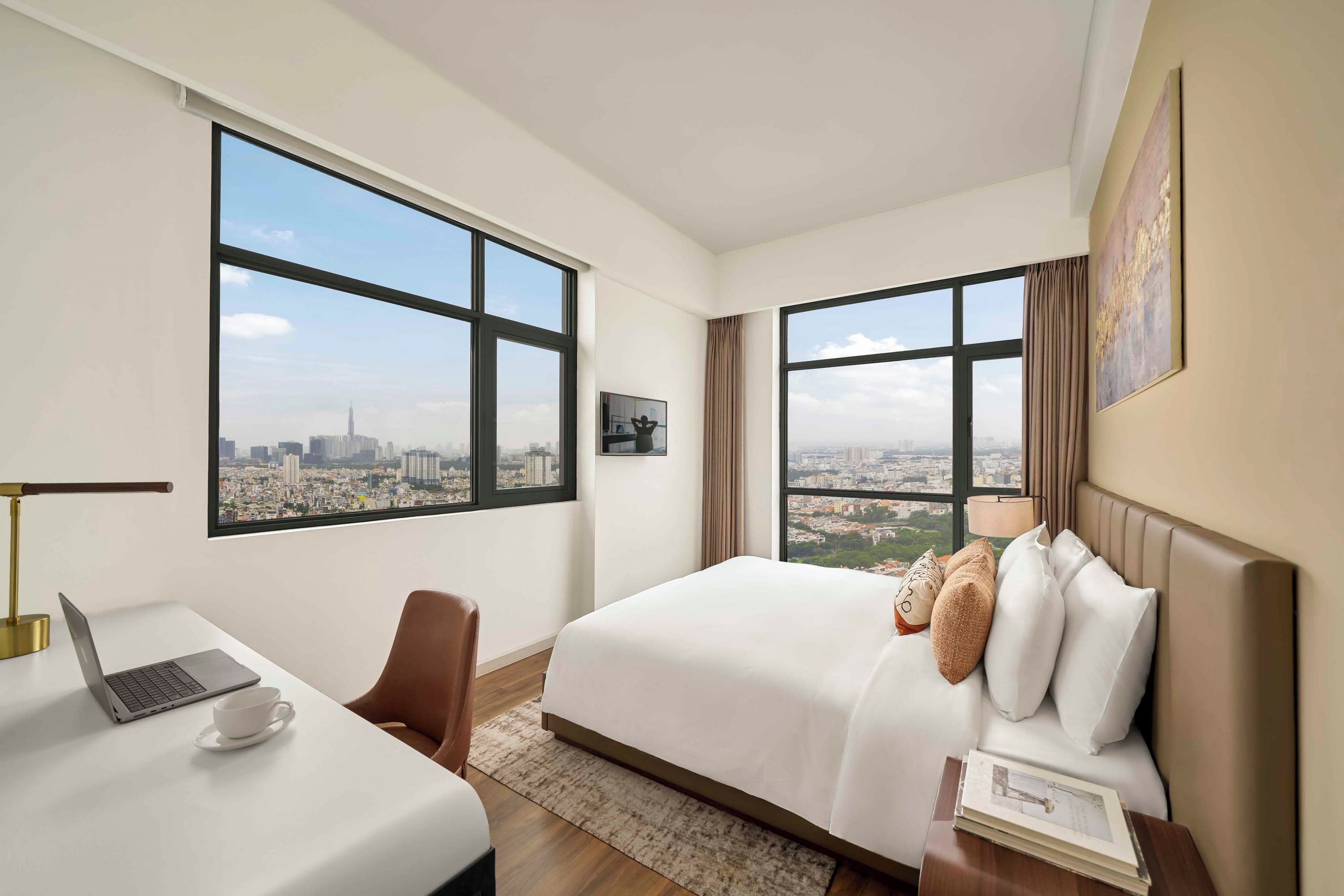 Una habitación de hotel con una cama, un escritorio con una computadora portátil y vistas a un paisaje urbano.