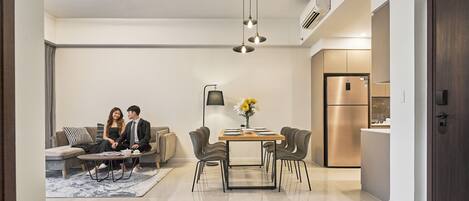 A modern living room with a sofa, dining table, and chairs. There is a kitchen area with a refrigerator and an air conditioning unit.