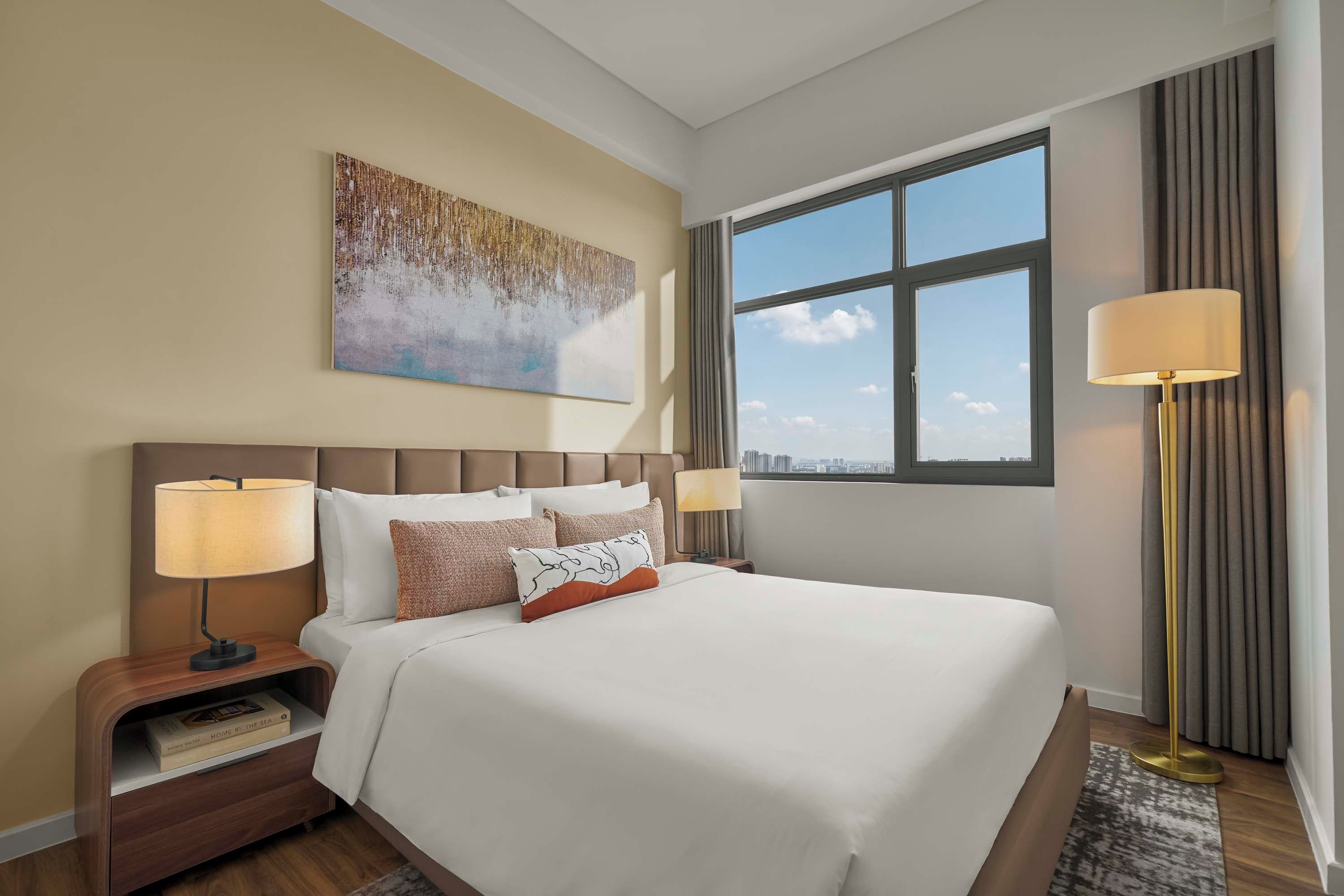 Quarto de hotel com uma cama grande, duas luminárias de cabeceira, mesa de cabeceira com um livro, um quadro na parede e uma janela com vista para a cidade.