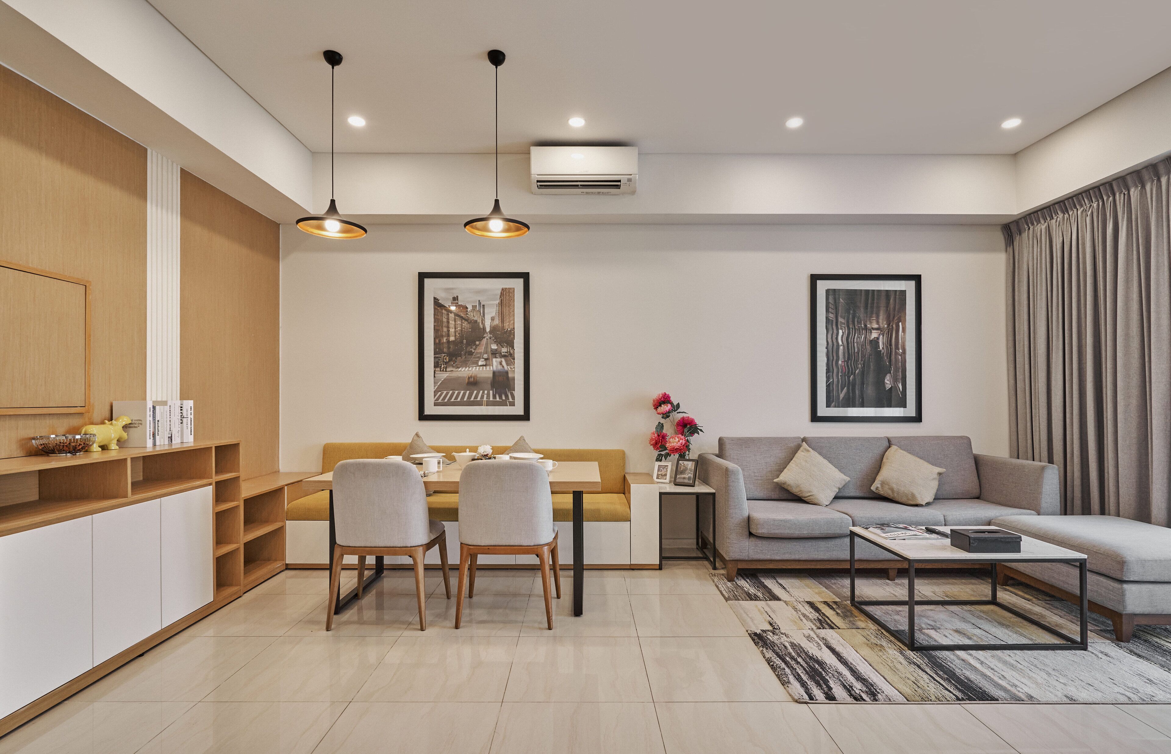 A modern living room with a sofa, dining table, and chairs. There are framed pictures on the walls, a small side table with flowers, and an air conditioning unit on the ceiling.