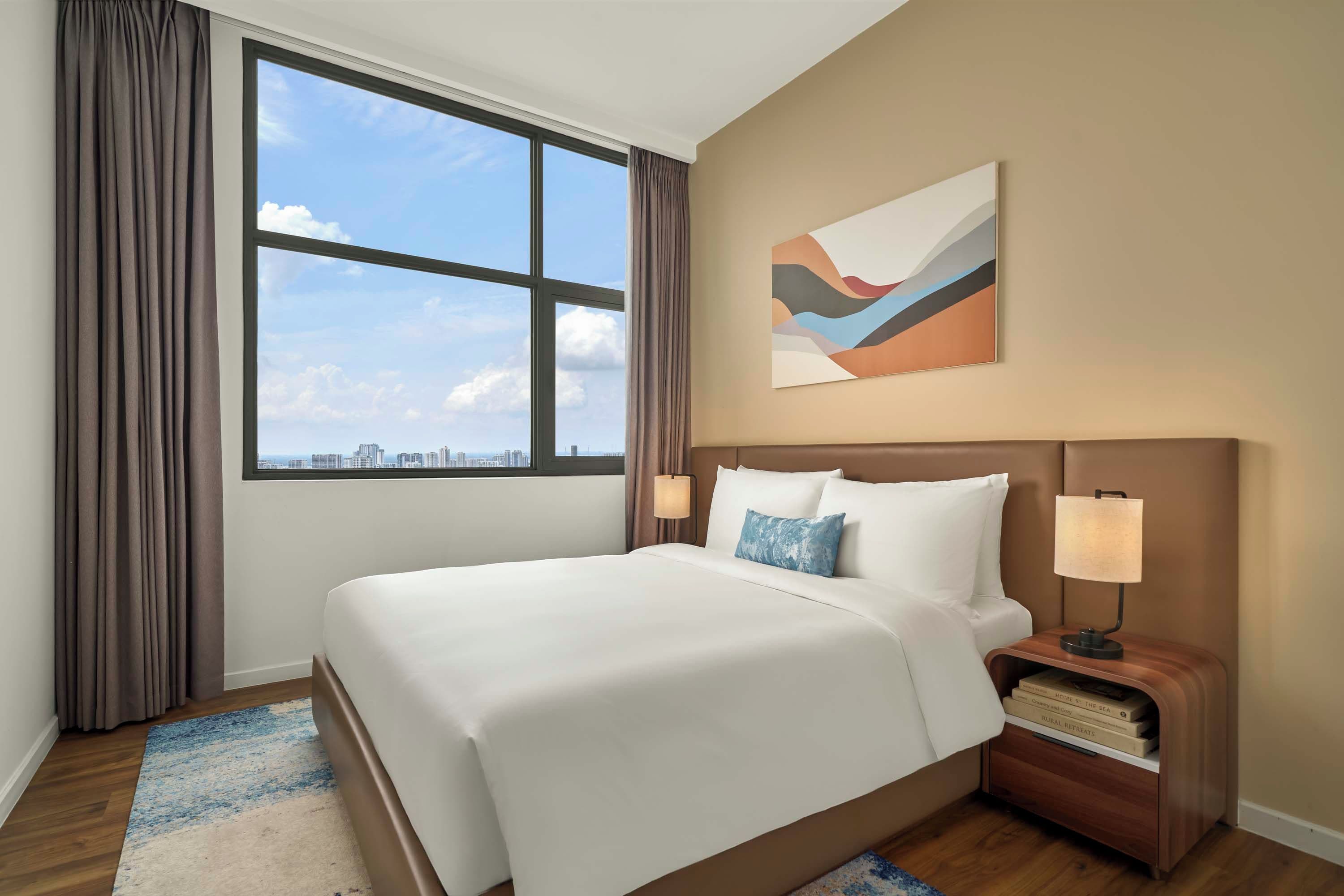 A hotel room with a large bed, a nightstand with a lamp, a city view, and a piece of abstract art on the wall.