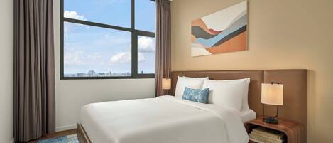 A hotel room with a large bed, a nightstand with a lamp, a city view, and a piece of abstract art on the wall.