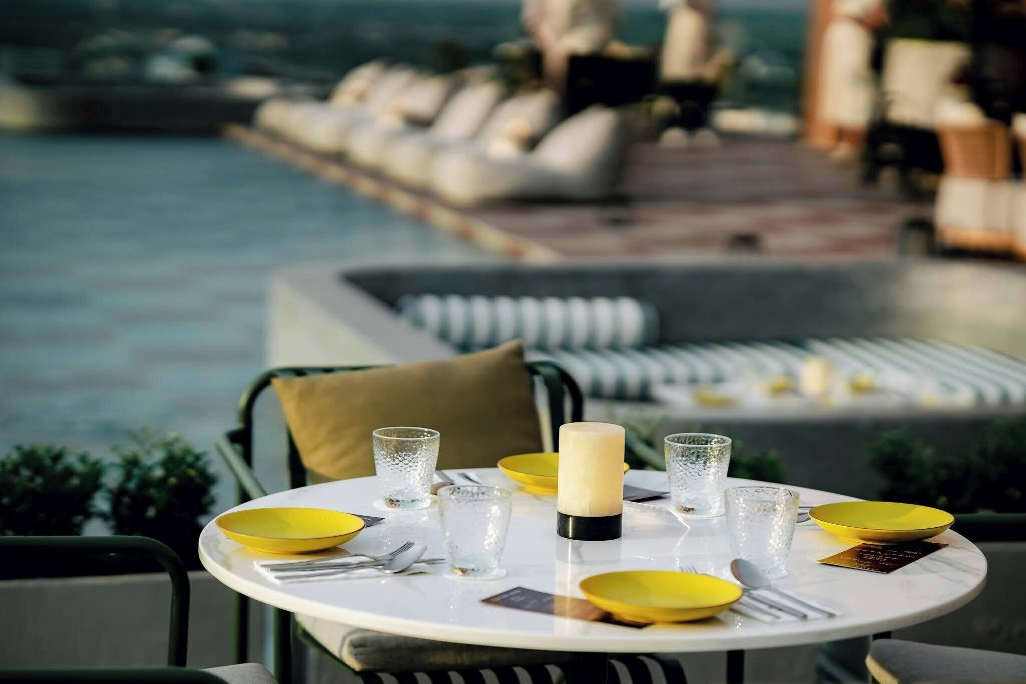 Une table à manger avec des assiettes jaunes, des verres transparents et une bougie, le tout disposé devant une vue sur la piscine.