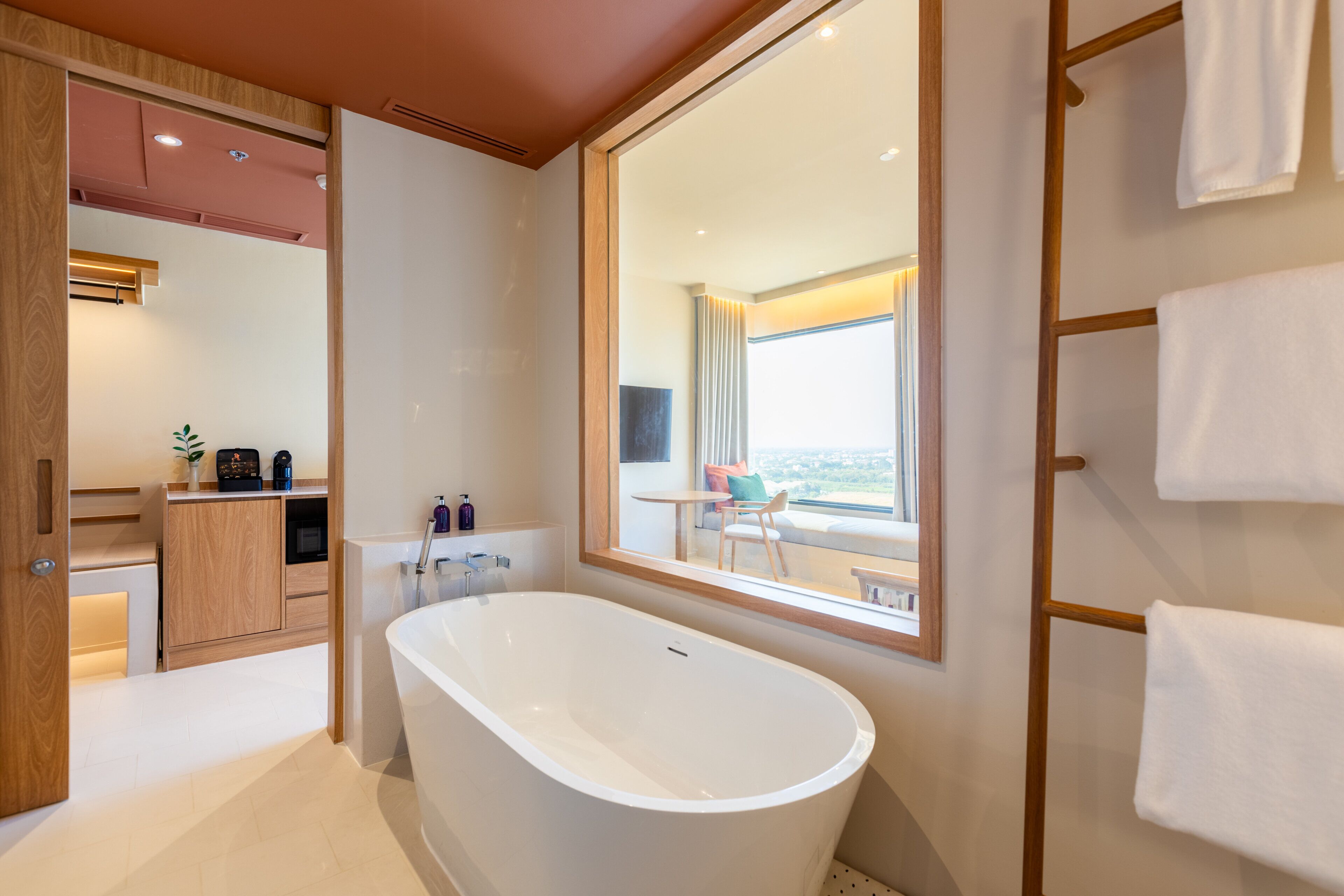 A modern bathroom with a freestanding bathtub, wooden cabinetry, and a large mirror reflecting a balcony with a view.