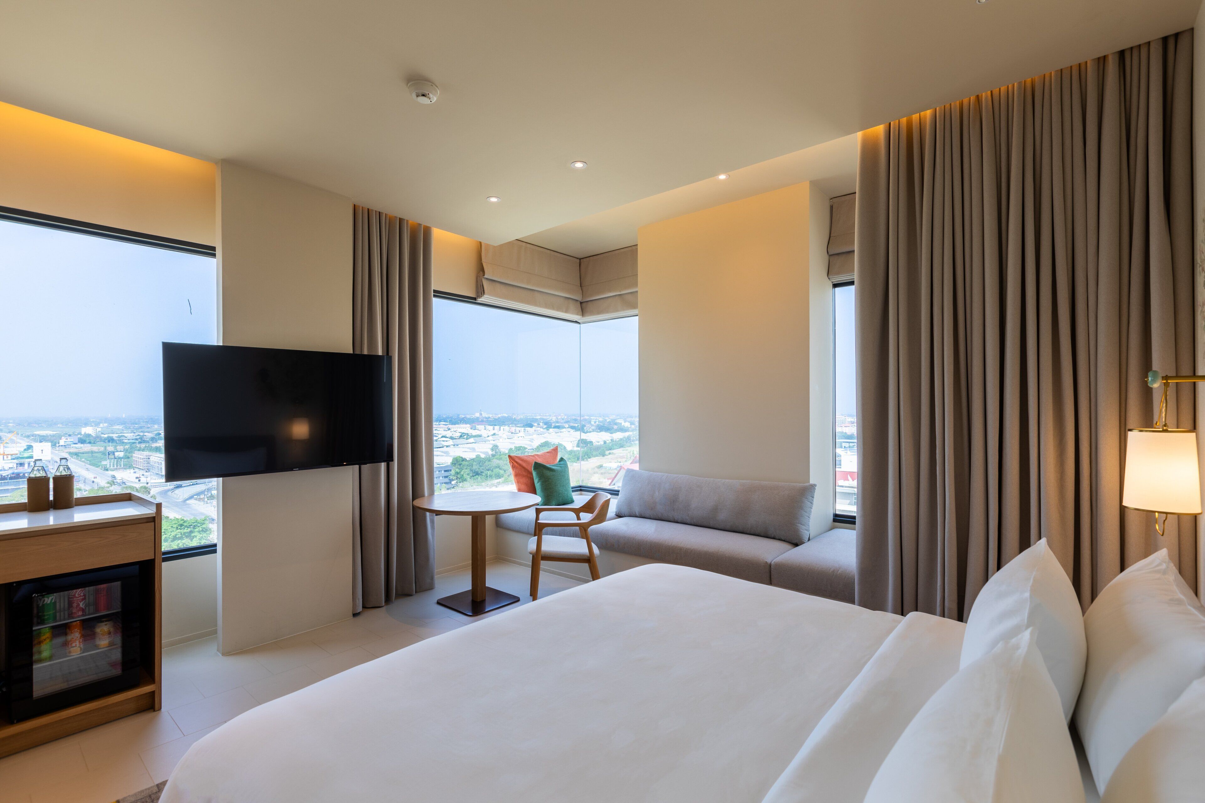 A modern hotel room with a large bed, a flat-screen TV, a small table with a chair, a sofa, and a view of the city through large windows.