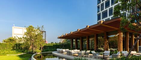 A modern outdoor lounge area with seating and a water feature, surrounded by greenery and a building in the background.