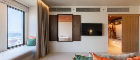 A modern hotel room with a large window, a sofa with cushions, a flat-screen TV, a mirror, a wardrobe, and a bathroom visible through an open door.