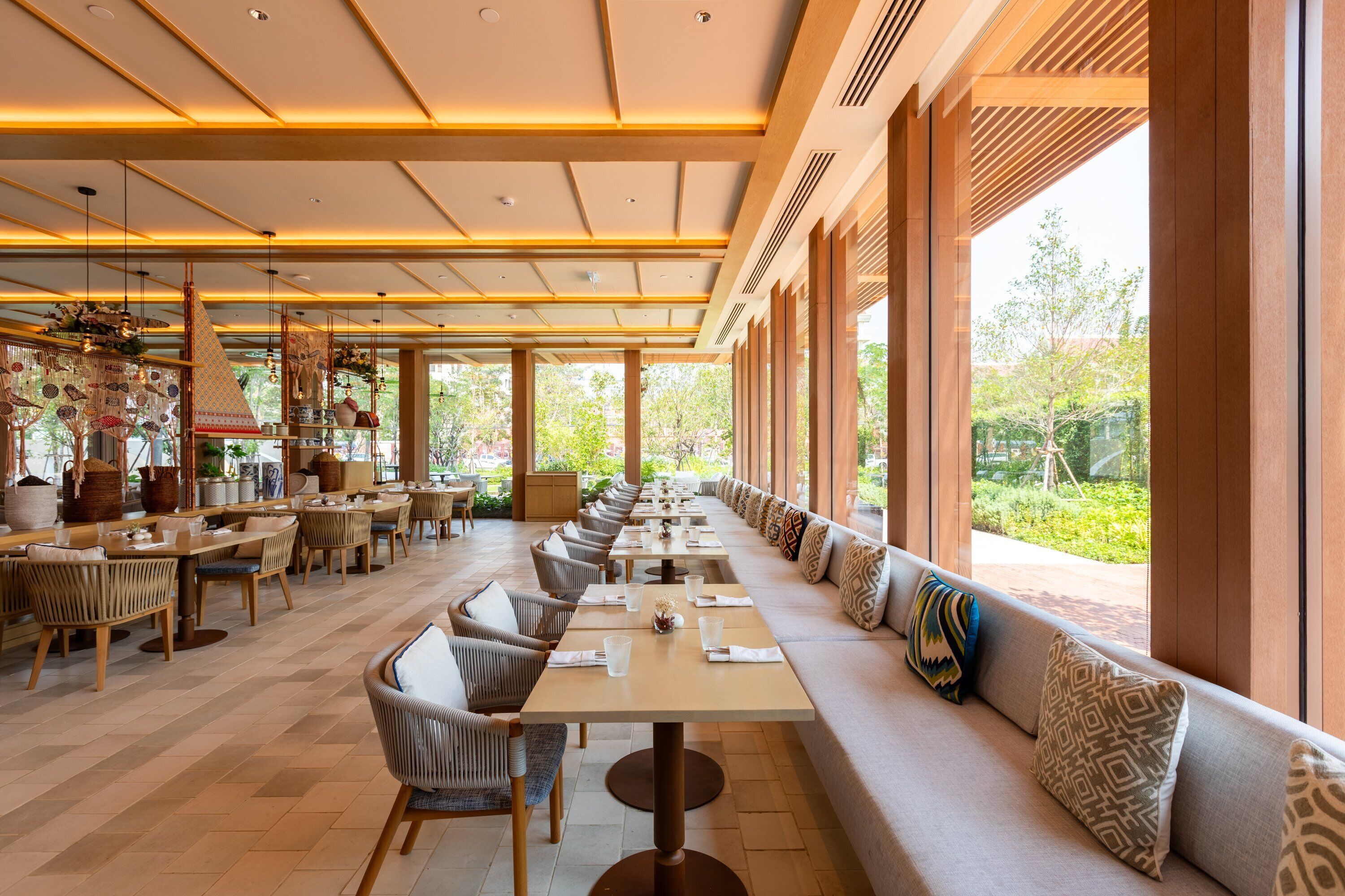 A spacious indoor dining area with long tables and comfortable seating, large windows offering a view of greenery outside.