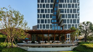 Un hotel moderno con una estructura de varios pisos, zona de estar al aire libre con sillas y mesas, y una pequeña fuente en la parte delantera.