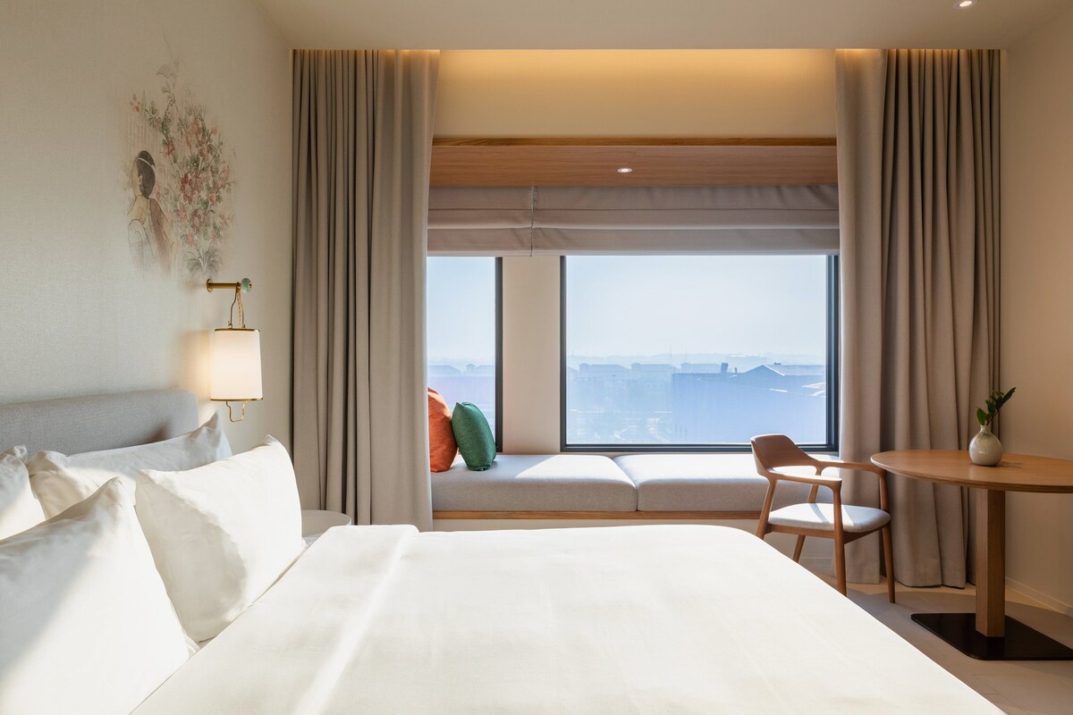A hotel room with a large bed, a bedside table, a chair, a small table, and a window with a city view.