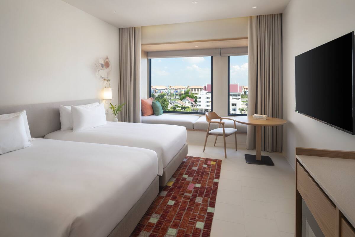 Een hotelkamer met twee bedden, een bureau, een televisie en uitzicht op gebouwen door grote ramen.