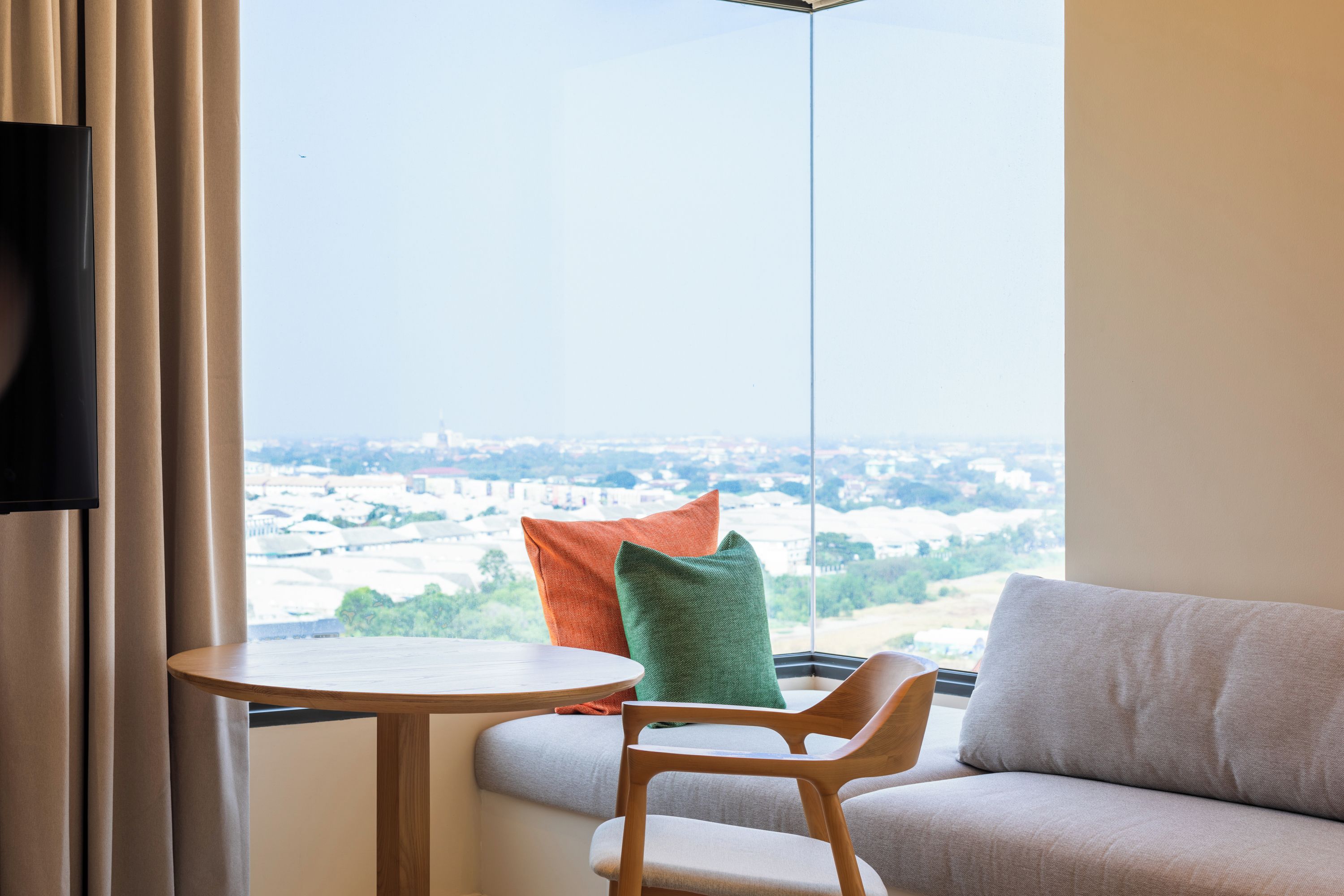 Una habitación con vistas a un paisaje urbano, una mesa redonda de madera, un sofá gris y cojines de colores.