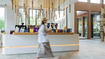 A modern hotel lobby with a reception desk, hanging pendant lights, and a view of greenery outside.