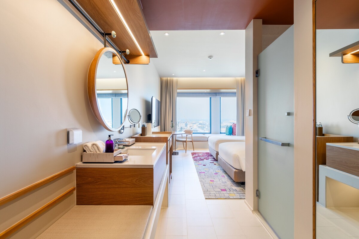 A modern hotel room with a large mirror, a wooden vanity, a bed, a small table with a chair, and a view of the city through large windows.