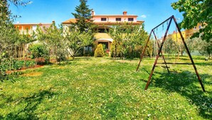 A large, two-story house with a red-tiled roof, surrounded by a well-maintained garden with various plants and a swing set.