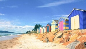 A row of colorful beach huts along a sandy beach with large rocks in the foreground and a clear blue sky. - By the Bay - 4pm check out Sundays* (Rosebud)