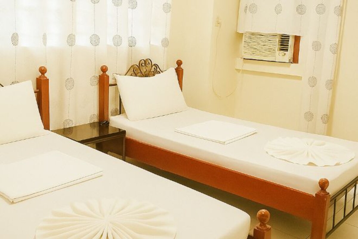 Une chambre d’hôtel avec deux lits simples, une table de chevet, une petite table d’appoint, un climatiseur mural et des rideaux à motifs.