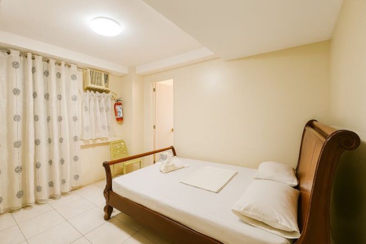 Une chambre à coucher avec un lit en bois, du linge de lit blanc, une fenêtre avec des rideaux, une table de chevet et un extincteur fixé au mur.