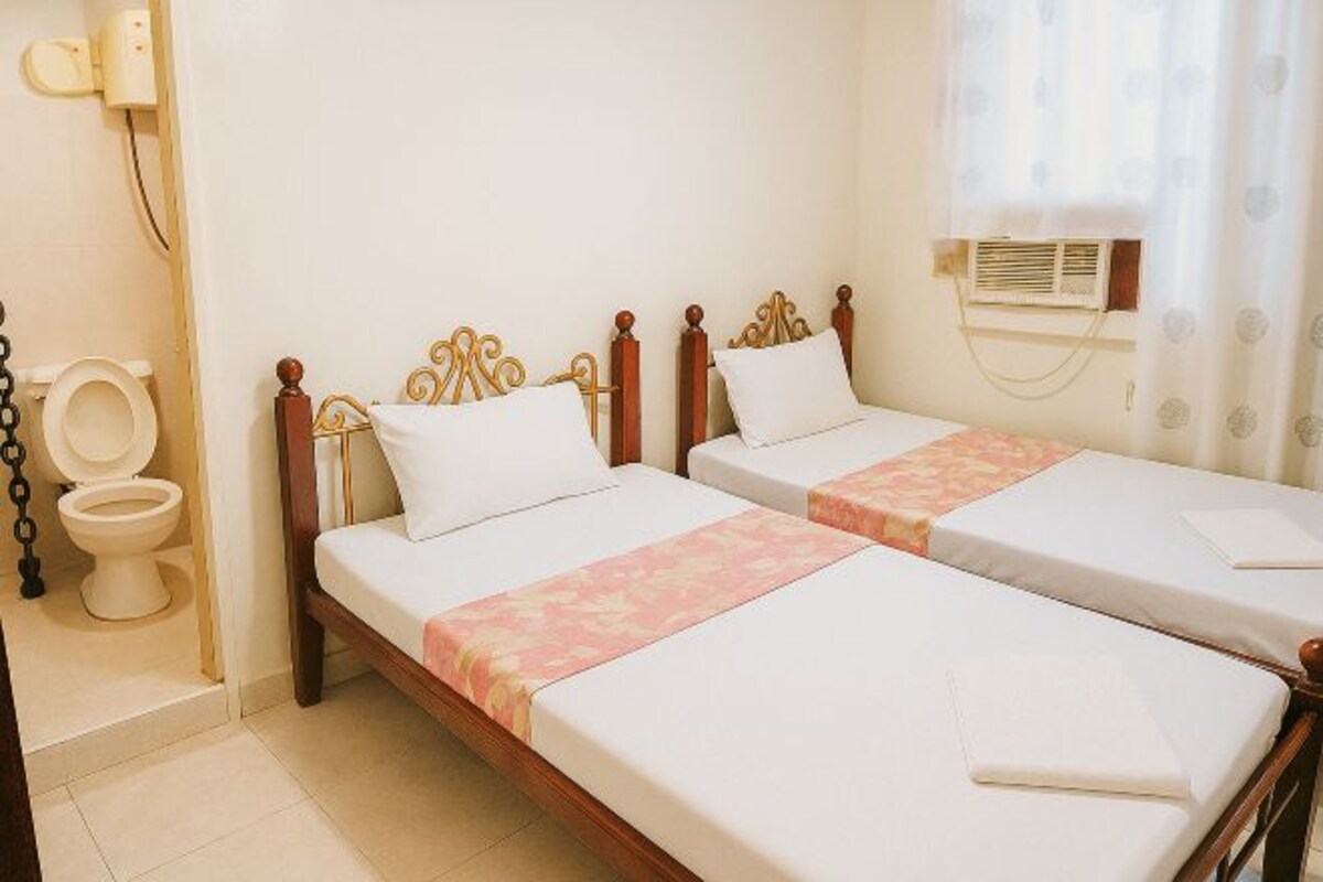 Une chambre d’hôtel avec deux lits simples, des toilettes et une fenêtre avec un appareil de climatisation.