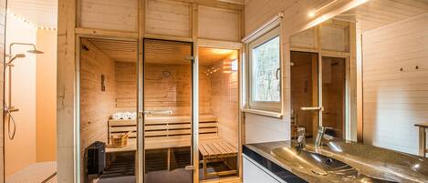 Una sala de sauna con bancos de madera y una puerta de vidrio, un lavamanos moderno y una ventana con vista a árboles.