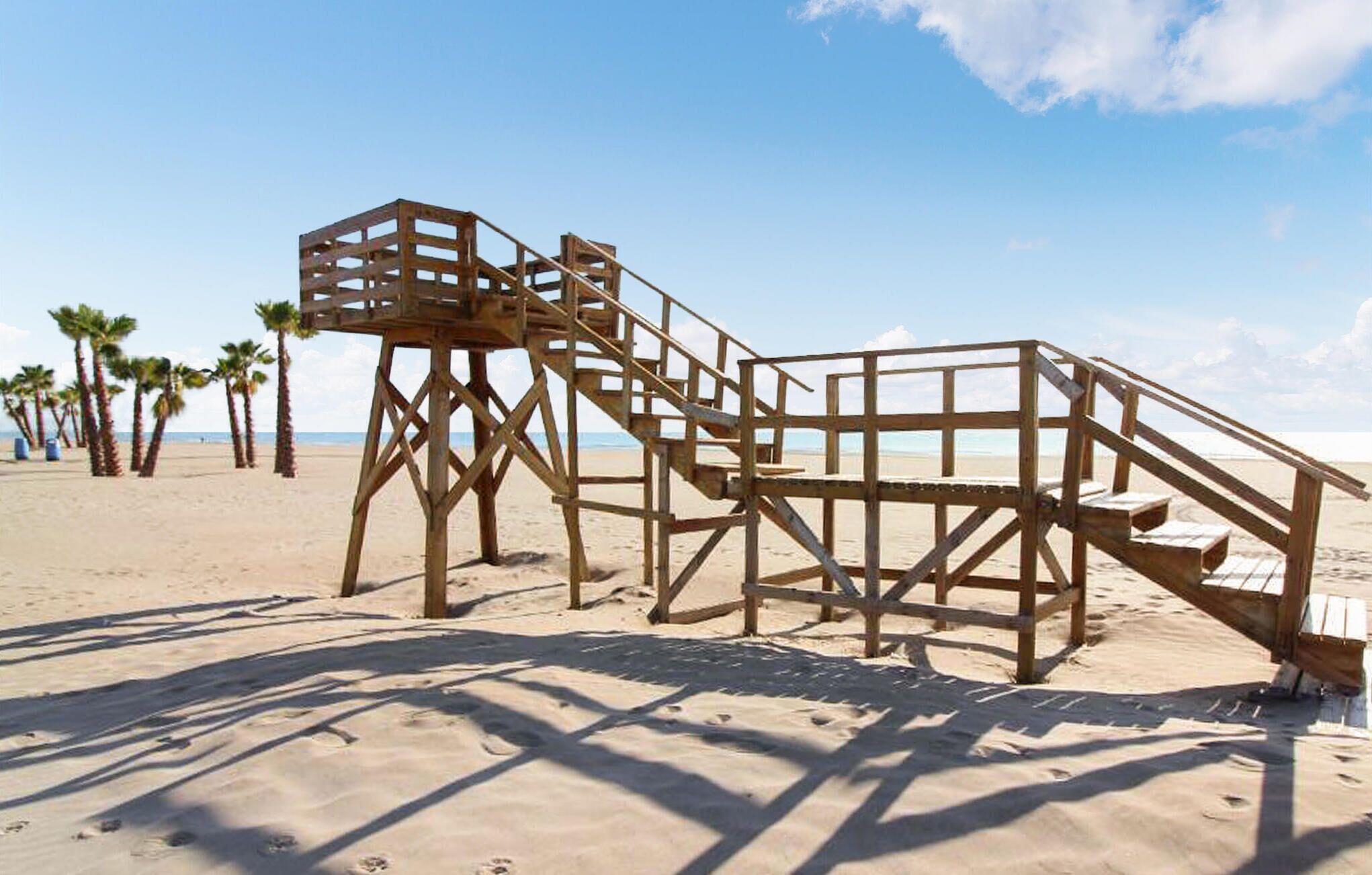 Una torre de salvavidas de madera en una playa arenosa con palmeras al fondo.
