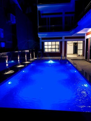Piscina com um recurso aquático iluminado em azul, rodeada por um edifício com grandes janelas e varanda.