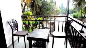 Un balcon doté d’une table en bois et de deux chaises, entouré d’une rambarde en métal, donnant sur un jardin où poussent diverses plantes et arbres.