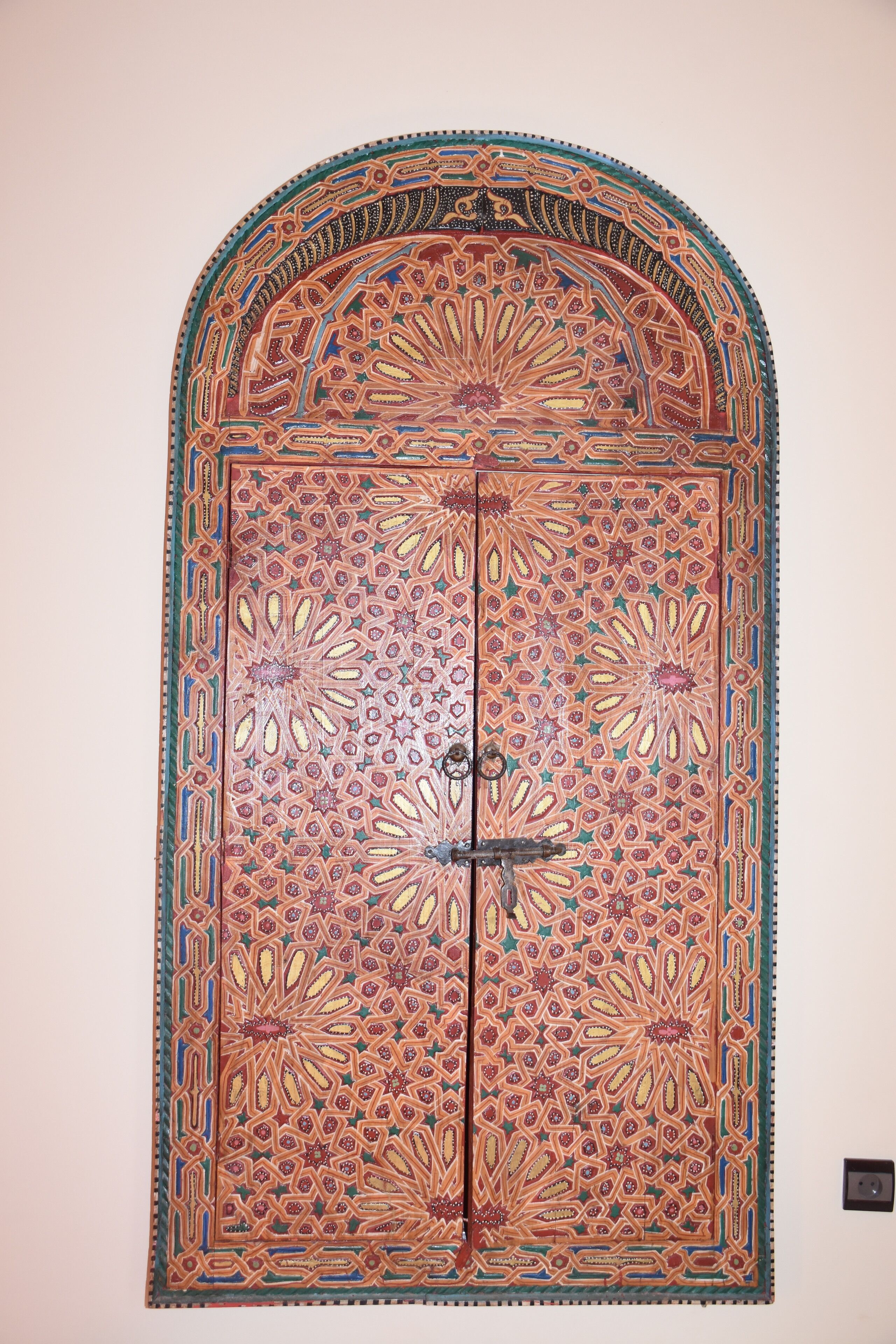 Une grande porte richement décorée, ornée de motifs géométriques et floraux complexes, dotée d’une poignée centrale et d’une arche décorative au sommet.