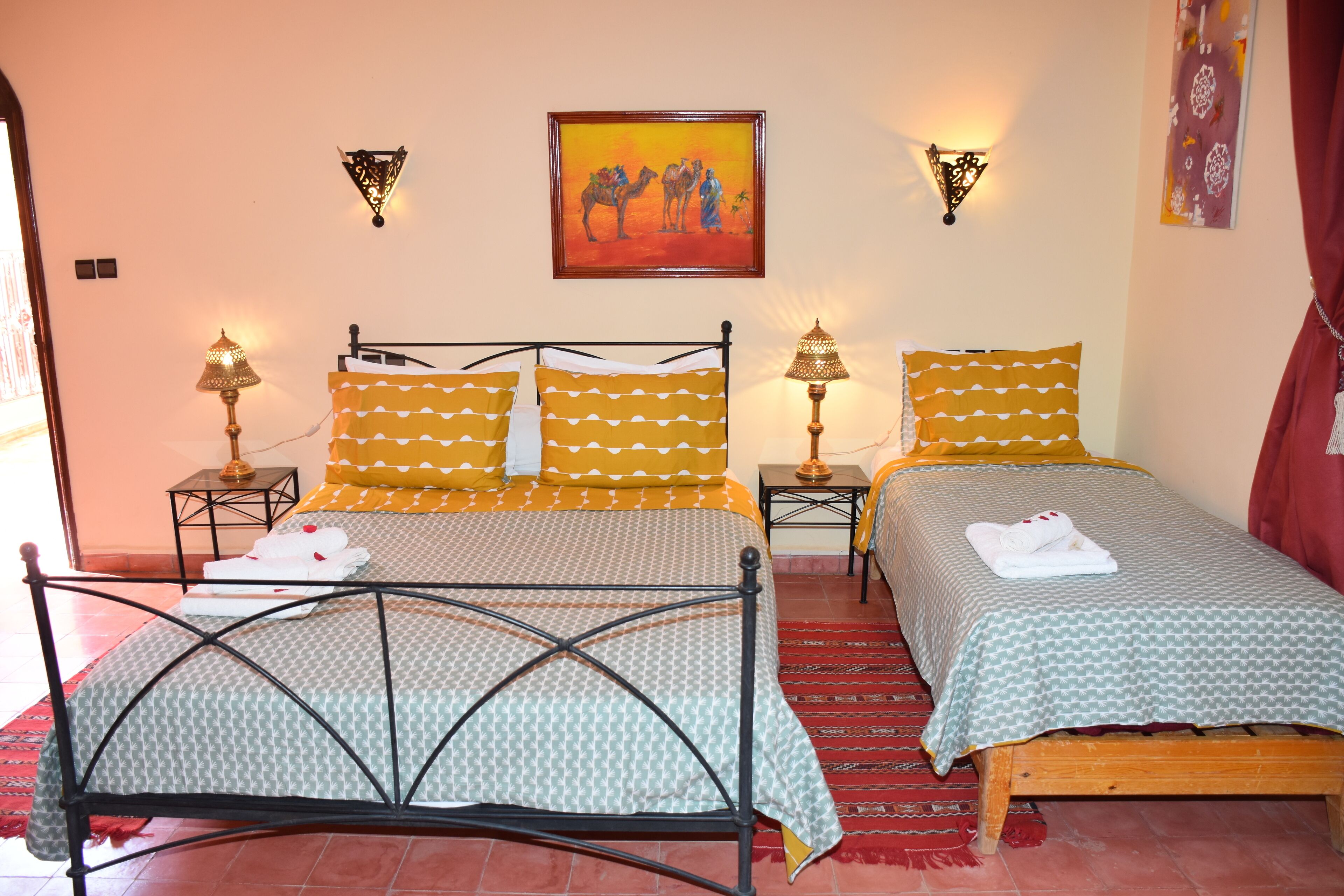 A double bed with a metal frame, two bedside tables with lamps, and a painting of camels on the wall.