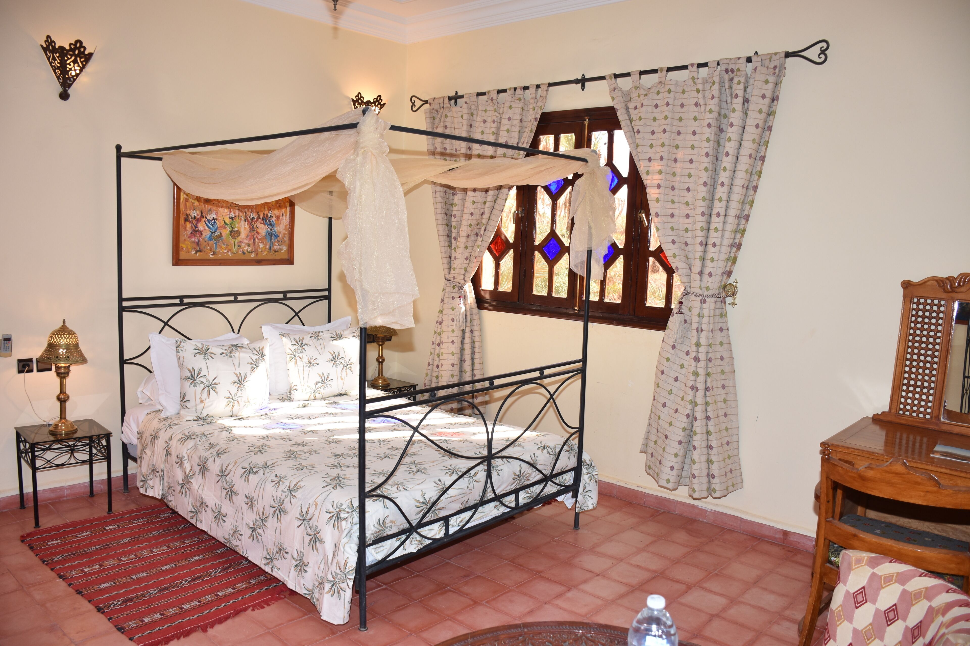 Una cama con dosel, una mesita de noche pequeña con una lámpara, una alfombra roja y un mueble auxiliar de madera.