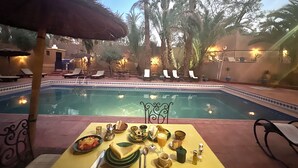 A poolside dining area with a table set for two, overlooking a swimming pool surrounded by palm trees and lounge chairs. - Riad le Petit Jardin  (Zagora)