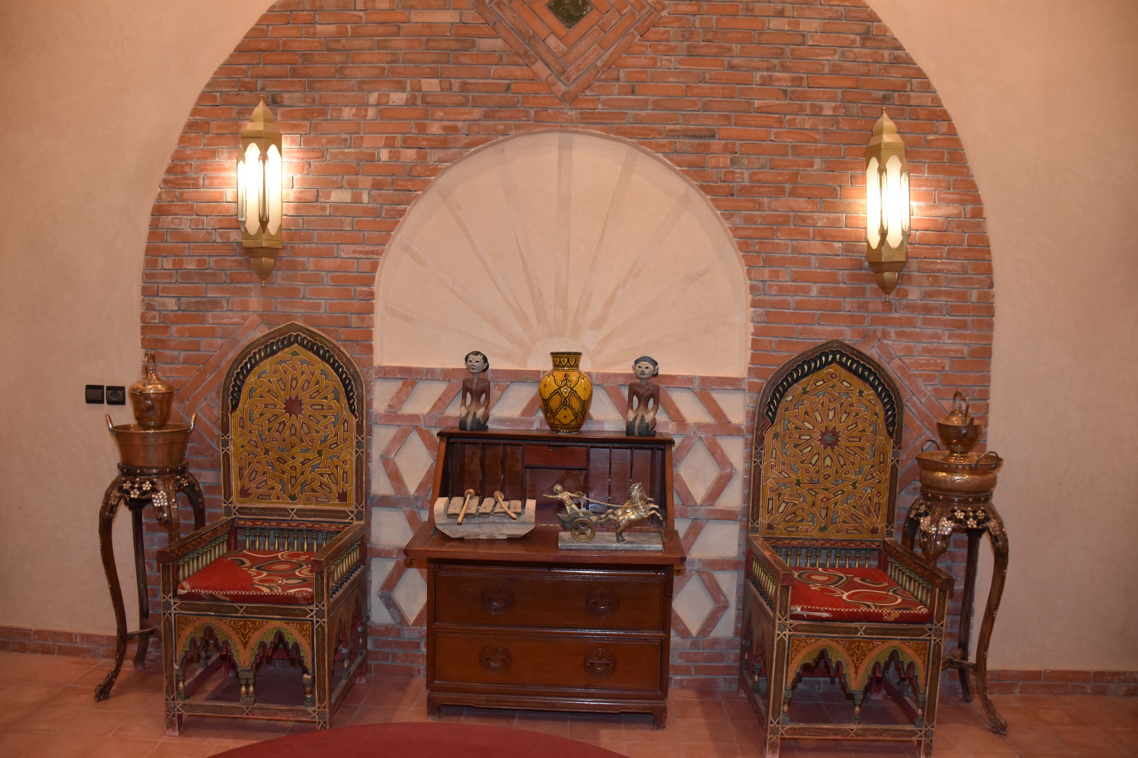 A room with two ornate chairs, a wooden chest of drawers, and a decorative wall with a fan.