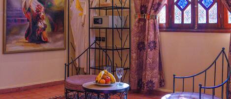 A room with a red carpet, a painting, a metal chair, a table with fruit and a glass, and a window with curtains.